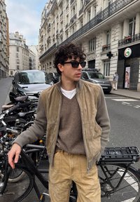 Νέος άνδρας με γυαλιά ηλίου, μπεζ μπουφάν και παντελόνια, στέκεται δίπλα σε ποδήλατα σε δρόμο της πόλης με ιστορικά κτίρια.