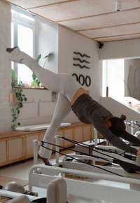 Biely Pilates reformer s čiernymi odporovými pásmi. Osoba v sivej košeli a bielych legínach vykonáva cvičenie. Prírodné svetlo z okien.