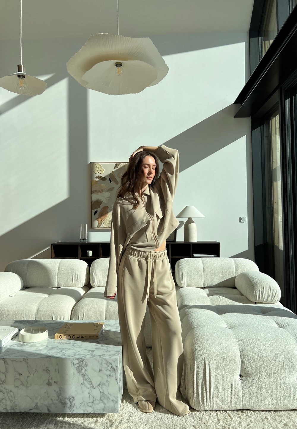 Beige loungewear set featuring an oversized top and drawstring pants, complemented by a textured cream sofa, marble coffee table, and pendant lights.