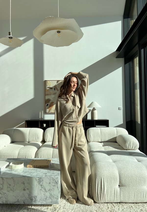 Beige lounge-set med oversized topp och dragkedjebyxor, kombinerat med en texturerad krämfärgad soffa, marmor kaffebord och pendellampor.
