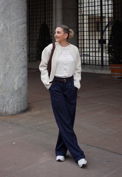 Chaqueta bomber texturizada blanca, camisa beige, pantalones anchos de rayas azules, cinturón marrón, zapatillas blancas. De pie junto a un pilar de piedra.
