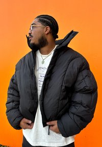 Crna pufasta jakna s krojenjem iznad struka, teksturiranom tkaninom, velikim napučenim rukavima i visokim ovratnikom, nosi se preko bijele majice s grafičkim printom.