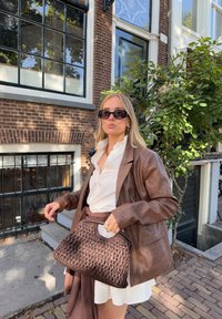 Bruine leren blazer over een witte blouse, met een donkerbruine gevlochten handtas met een gebogen handvat. De achtergrond bestaat uit een bakstenen muur en loof.