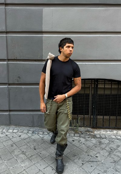 Camiseta negra ajustada, pantalones cargo verde oliva con bolsillos laterales, botas negras hasta la rodilla y una bufanda beige texturizada caída sobre el hombro.