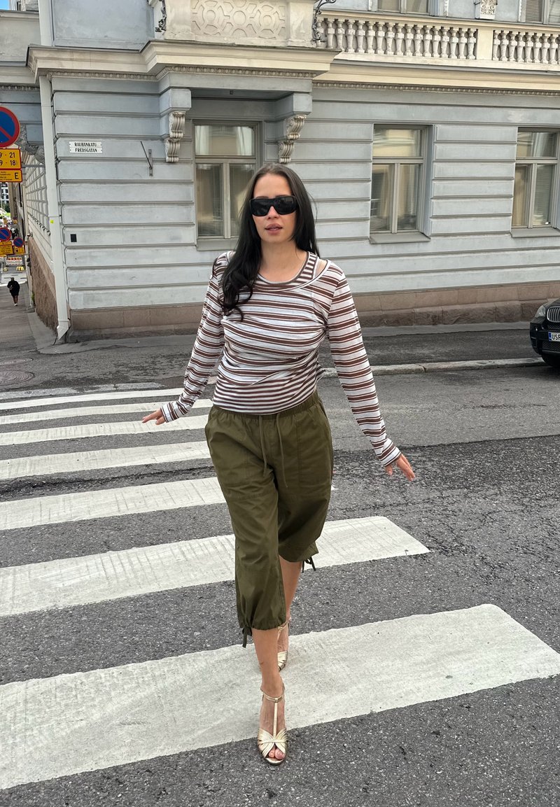 Femme portant des lunettes de soleil, une chemise à manches longues rayée, un pantalon olive et des sandales, traversant un passage piéton en ville.