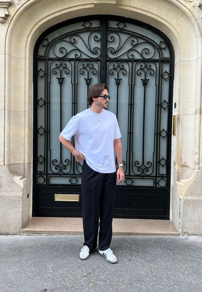 Camiseta blanca de manga corta, pantalones anchos negros y zapatillas blancas. Los accesorios incluyen gafas de sol y un collar de plata. Detrás hay una puerta negra ornamentada.