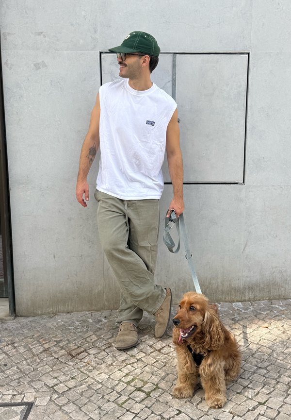 Homme portant une casquette verte et un débardeur blanc s'appuie contre un mur en tenant une laisse attachée à un chien assis de couleur brun doré.