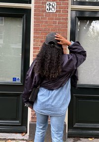 Zwarte satijnen bomberjack over een lichtblauw denimshirt, lichtblauwe jeans en een donkere pet, staand voor een bakstenen muur.