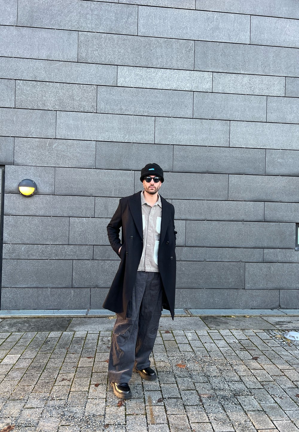 Cappotto doppiopetto nero su una camicia a fantasia, abbinato a pantaloni larghi scuri e stivali neri. Gli accessori includono un berretto e occhiali da sole.