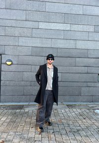 Cappotto doppiopetto nero su una camicia a fantasia, abbinato a pantaloni larghi scuri e stivali neri. Gli accessori includono un berretto e occhiali da sole.