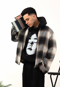 Jeune homme aux cheveux courts et bruns tenant un boombox vintage sur son épaule, portant une veste à carreaux et un sweat à capuche avec un imprimé de visage.