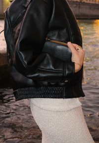 Fekete bőr bombázó dzseki, textúrázott felülettel, ráncolt derékrésszel és cipzáras zsebbel. Krémszínű pulóver anyag alatta, ami harmonizál a sötét háttérrel.