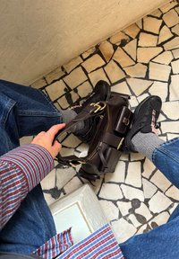 Brun lædertaske med guldfarvet hardware, omgivet af mørke sneakers og grå sokker, på et tekstureret, mønstret flisegulv. Stribet skjorte synlig.