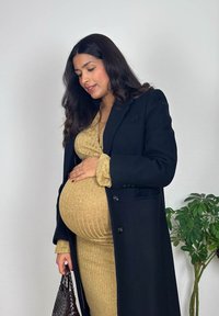 Femme enceinte en robe côtelée beige et manteau noir tenant doucement son ventre, portant un sac à main en osier marron, debout près d'une plante verte en pot.