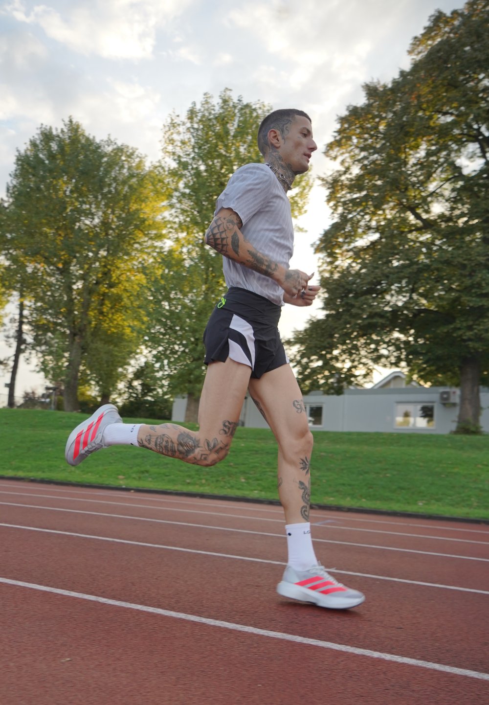 Corridore con tatuaggi che indossa pantaloni corti neri, maglietta bianca e scarpe da ginnastica bianche con strisce rosse che corre su una pista all'aperto in un parco.