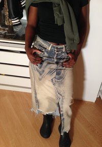 Personne portant un haut noir, une jupe en denim délavé usée, des chaussures Crocs noires, et un sweat-shirt vert drapé sur les épaules à l'intérieur.