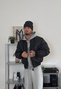 Mustast bomber-jakk ees on lukuga ees, soonikute ja kraegaga, halli kapuutsiga sviitri peal, kantud koos hallide treeningpükstega.