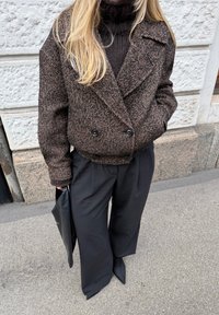 Άτομο με καφέ σακάκι διπλής οσμής σε σχέδιο ψαροκόκκαλο, μαύρο ζιβάγκο, φαρδιά μαύρη παντελόνια, κρατώντας μαύρο φάκελο, στέκεται στο πεζοδρόμιο.