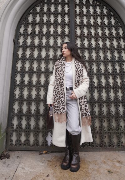 Bufanda de estampado de leopardo, abrigo crema, top blanco, pantalones grises, botas marrones hasta la rodilla; modelo posando frente a una puerta de hierro decorativa.