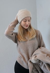 Bēšas krāsas ribota trikotāžas cepure un mīksts bēša džemperis ar garām piedurknēm, turēts kopā ar gaiši brūnu mēteli, uz gaiši pelēka fona.