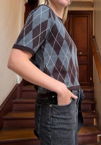 Άτομο που φοράει καφέ και γκρι πουλόβερ με μοτίβο αργκάιλ κοντομάνικο και σκούρο γκρι τζιν, με το ένα χέρι στην τσέπη, στέκεται δίπλα σε ξύλινη σκάλα.