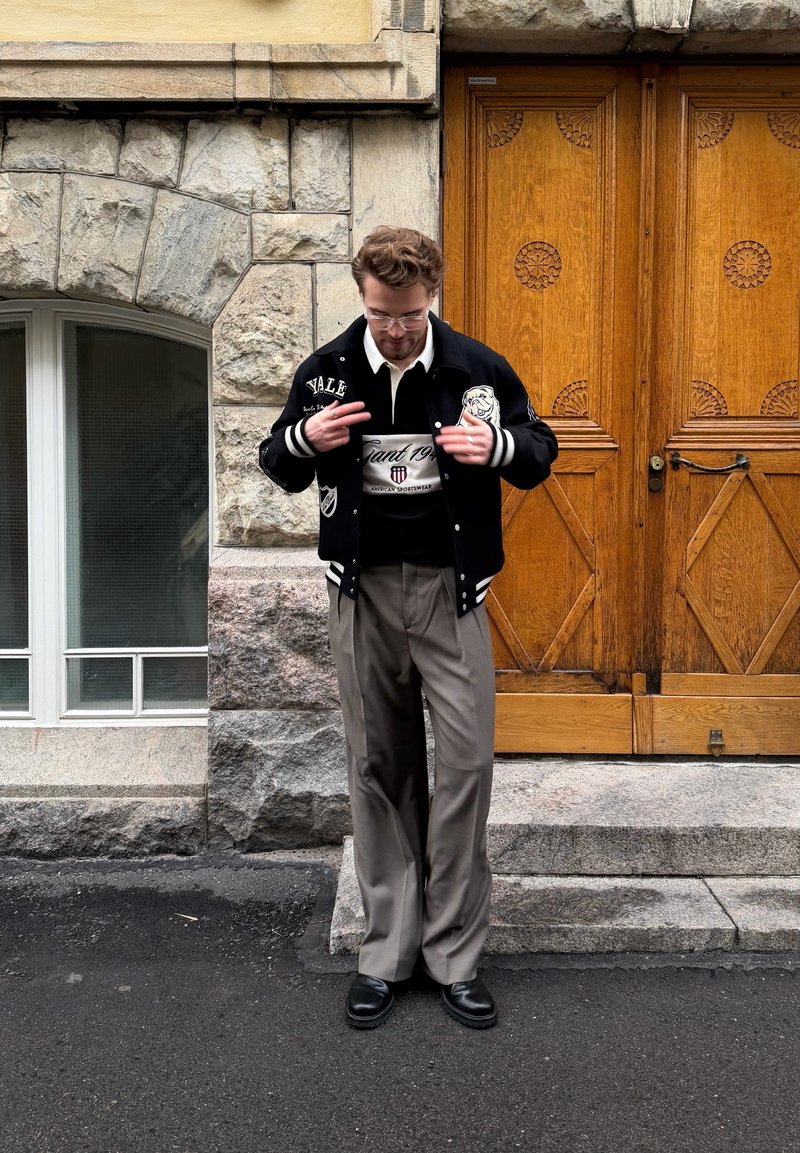 Homme portant une veste universitaire noire et un pantalon beige, debout sur des marches en pierre devant des portes doubles en bois ornées et un mur en pierre.