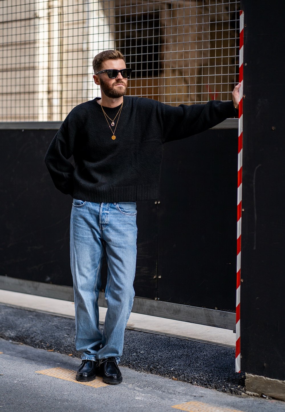 Suéter negro de gran tamaño, jeans rectos de mezclilla azul claro, zapatos negros. Los accesorios incluyen collares de oro en capas y gafas de sol.
