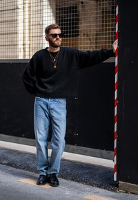 Suéter negro de gran tamaño, jeans rectos de mezclilla azul claro, zapatos negros. Los accesorios incluyen collares de oro en capas y gafas de sol.