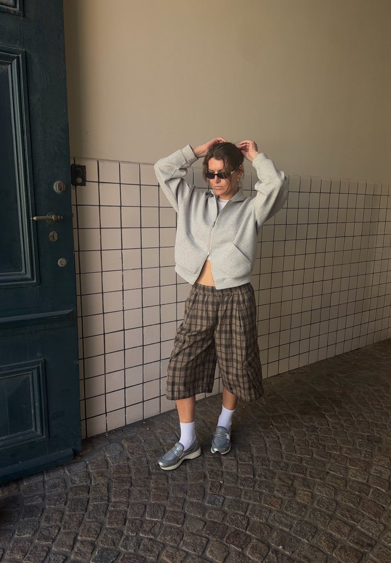 Άτομο με γυαλιά ηλίου, γκρι ζακέτα με φερμουάρ, καρό σορτς μέχρι το γόνατο, λευκές κάλτσες και ασημένια παπούτσια που στέκεται σε πλακόστρωτο δάπεδο κοντά σε πλακιδωμένο τοίχο.