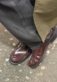 Mocasini maro din piele cu finisaj lucios, având cusături decorative și vârf rotund, asortați cu pantaloni din denim închis la culoare.