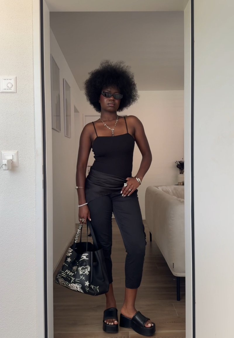 Sieviete ar afro frizūru, tērpusies melnā tanktopā, melnās biksēs, platformu sandales, saules brilles un tur melnu grafisko tote somu iekštelpās.