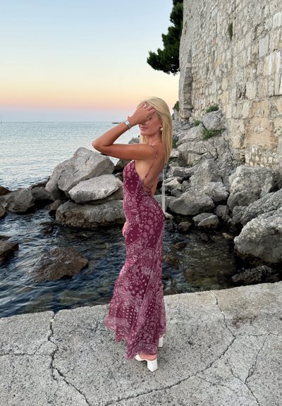 Vestido maxi en tela burdeos con un patrón de paisley, que presenta tirantes finos y un corte en la espalda. Combinado con tacones blancos de bloque.