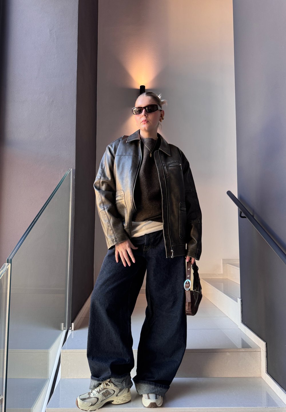 Casaco de couro preto sobre um sweater cinza, combinado com jeans de denim escuros e largos e tênis cinzas. Acessorado com óculos de sol e uma bolsa.