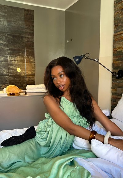 Un vestido verde y con volantes se despliega sobre una cama con almohadas blancas. El entorno presenta una pared de piedra, una lámpara negra y elementos decorativos sobre una superficie.