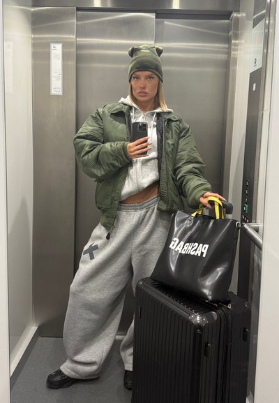 Chaqueta bomber verde sobre una sudadera gris con capucha, pantalones de chándal grises con un acento negro, una bolsa negra con el texto "PASHA" y una maleta negra en un ascensor.