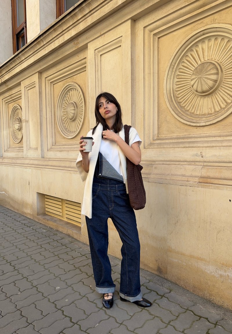 Jeune femme en tenue décontractée tenant une tasse de café, debout sur un trottoir pavé devant un bâtiment beige avec des motifs circulaires décoratifs.
