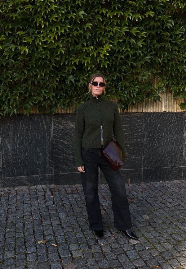 Femme portant un pull zippé vert foncé, un pantalon noir, des lunettes de soleil et tenant une grande pochette marron, debout sur un pavé à côté d’un mur couvert de lierre.