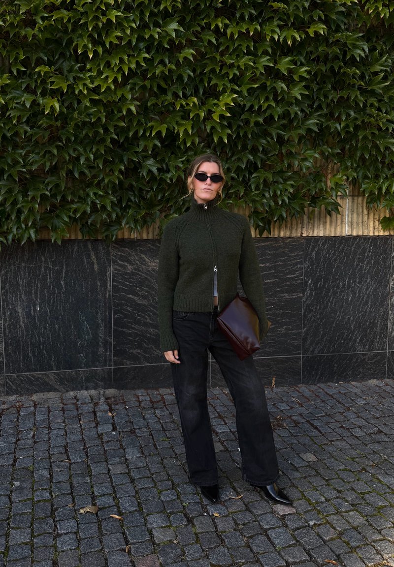 Femme portant un pull zippé vert foncé, un pantalon noir, des lunettes de soleil et tenant une grande pochette marron, debout sur un pavé à côté d’un mur couvert de lierre.