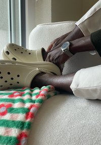 Személy bézs Crocs cipőben és világos nadrágban, fehér texturált bútoron ülve, mellette színes csíkos takaró. Kezei összekulcsolva a lába felett.