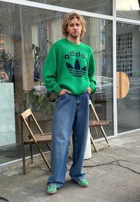 Πράσινο πλεχτό πουλόβερ με ναυτικό λογότυπο "adidas", σε συνδυασμό με χαλαρές μπλε τζιν και πράσινα παπούτσια slip-on, στέκεται μπροστά σε γυάλινο παράθυρο.