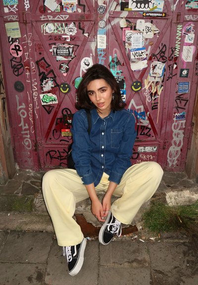 Camisa de mezclilla con botones, pantalones amarillos de pierna ancha y zapatillas negras. El fondo presenta una puerta roja texturizada cubierta de varias pegatinas y graffiti.