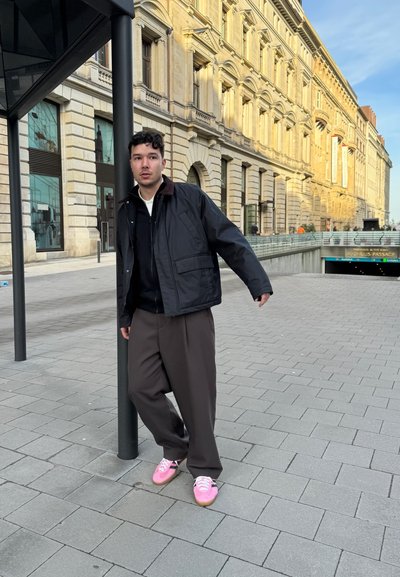 Chaqueta negra con cuello marrón, suéter negro debajo, pantalones marrones holgados y zapatillas rosas con acentos negros. Entorno urbano de día.