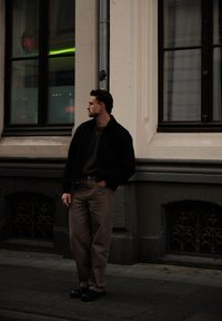Black jacket, dark green jumper, light beige trousers, and black shoes. Standing near a building with large windows and decorative metal accents.