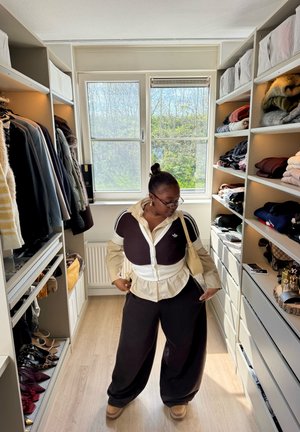 Persona con ropa beige y negra en capas de pie en un vestidor con estantes de ropa doblada y abrigos colgados junto a una ventana.