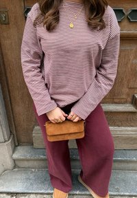 Femme assise sur des marches en pierre, portant une chemise à manches longues rayée bordeaux, un pantalon bordeaux, des bottes couleur chameau, tenant un petit sac à main en daim marron.