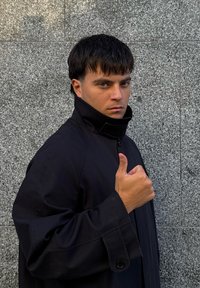 Fekete, magas galléros dzseki textúrált felülettel. A modell egy hüvelykujjfelnyújtós gesztust mutat a szürke kő háttér előtt.