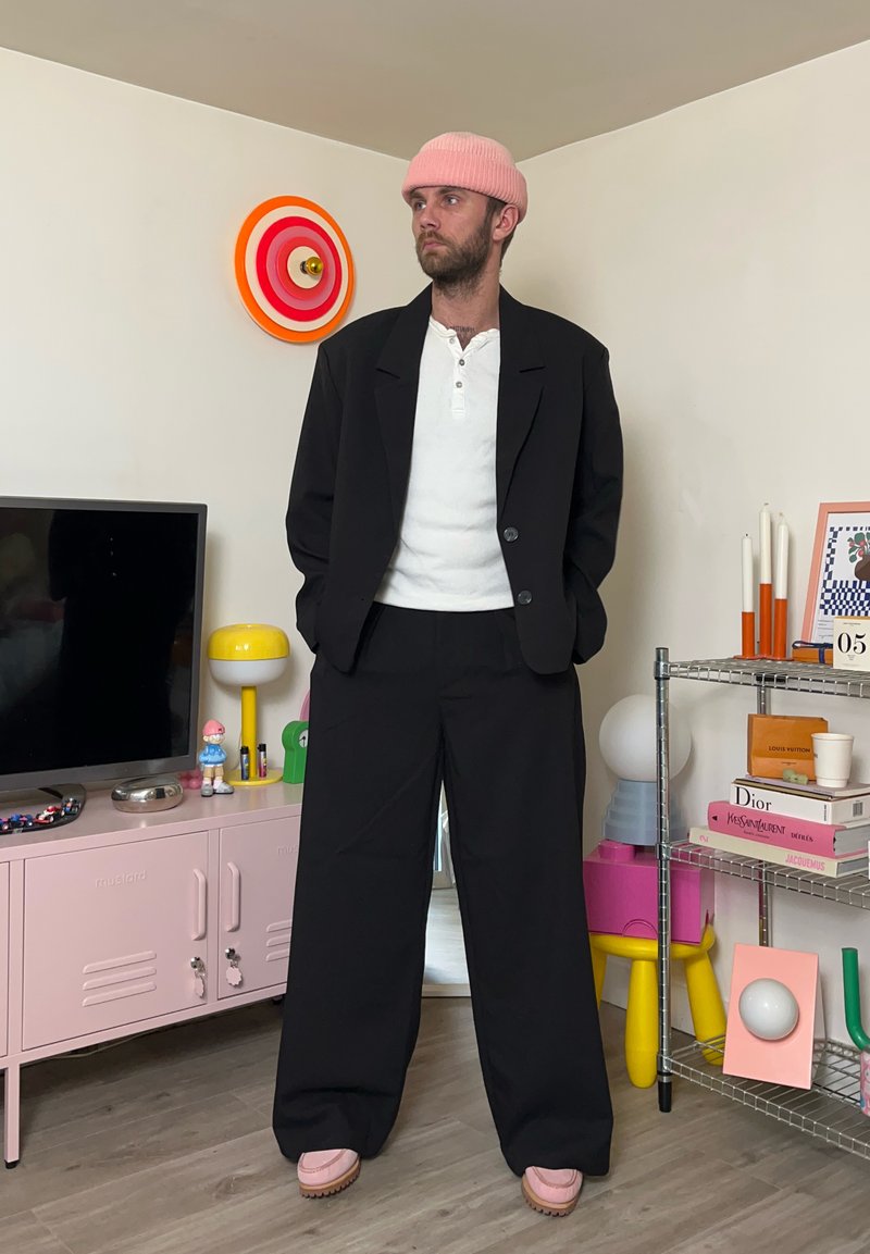 Hombre con gorro rosa, camisa blanca, blazer negro oversize y pantalones, de pie en una sala de estar moderna y colorida.
