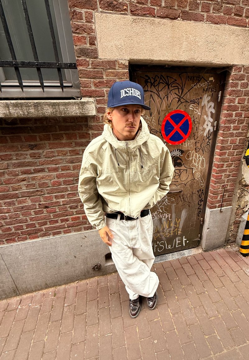 Lette beige jakke med høj krave, hvide oversized bukser og sorte sko. Mørkeblå kasket med hvidt logo. Murstensvæg i baggrunden.
