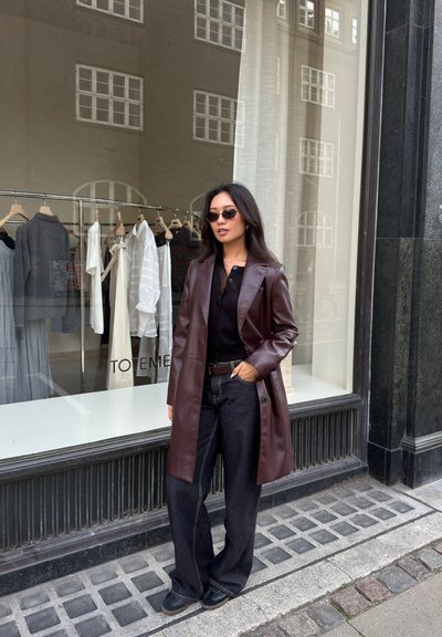 Largo abrigo de cuero burdeos, camiseta negra, pantalones oscuros de pierna ancha, zapatos negros y gafas de sol; diseño moderno exhibido junto a una vitrina.