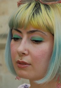 Ung person med hår til hagen, der har gule rødder og turkise spidser. Makeup inkluderer grøn øjenskygge med pink accenter. Guld piercinger.